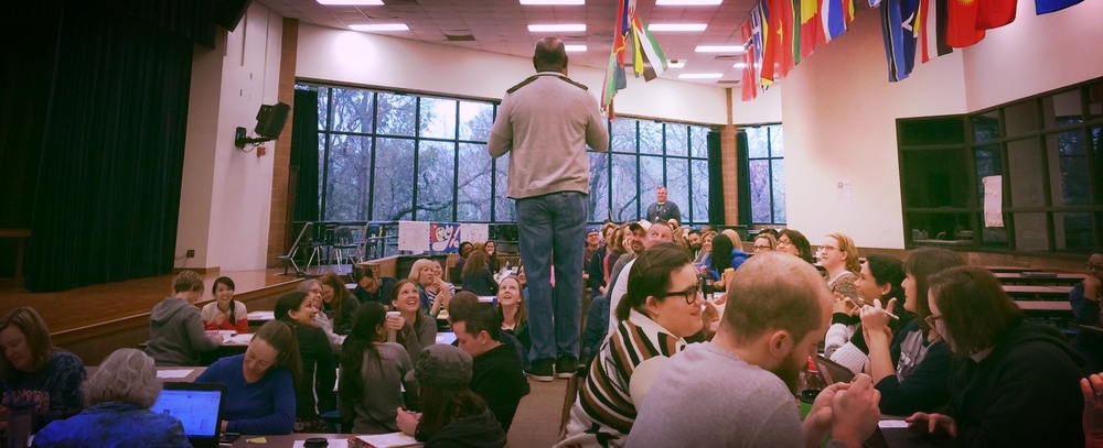 Teachers give attention to Assistant Principal Luiz Lazaro as he has fun speaking from the elevated platform in the cafeteria during a professional development day. Photo by Kara Wilkinson